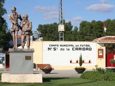 Esculturas de don Quijote y Sancho Panza ante la fachada blanca con el rótulo Campo Municipal de Fútbol Nuestra Señora de la Caridad.